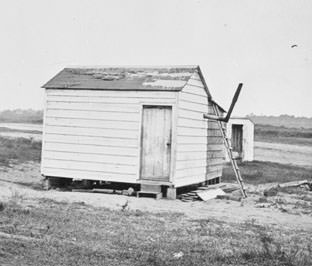 A simple Mitchelville house with a stove pipe. Notice the privy in the background.